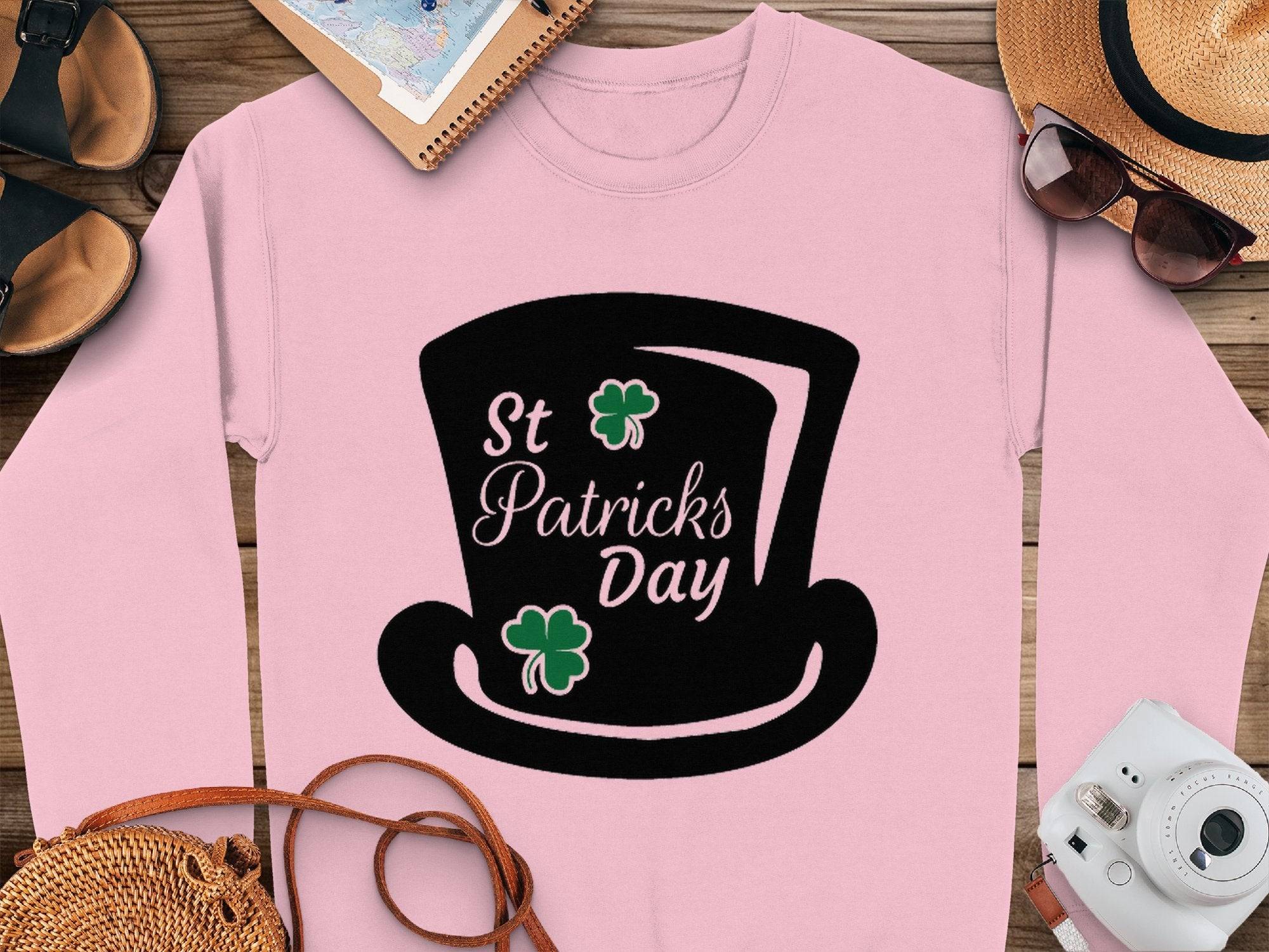 Garment Graphics presents a pink sweatshirt with a black top hat and green clovers graphic, featuring St Patricks Day text. It embodies festive spirit, displayed on wood and surrounded by accessories like a hat, sunglasses, shoes, notebook, and camera.