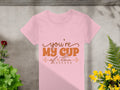 A pink 100% cotton T-shirt from Garment Graphics reads Youre My Cup of Tea in orange, with small decorative flourishes. Its displayed on a concrete surface, surrounded by green plants and yellow flowers.