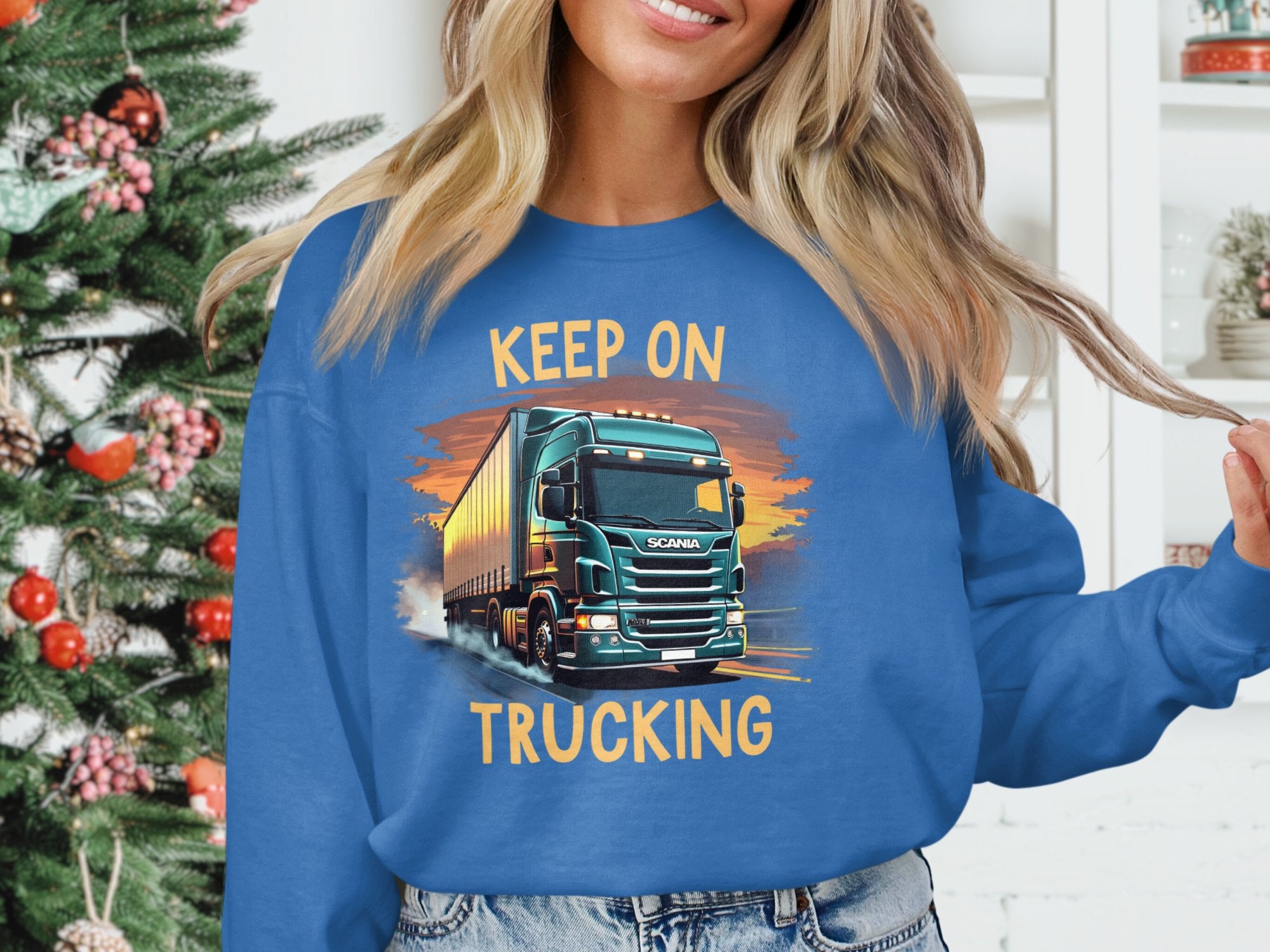 A truck enthusiast sports a Garment Graphics blue sweatshirt with a big rig and Keep On Trucking indoors by a festive Christmas tree.