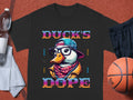 Garment Graphics Ducks Dope T-Shirt showcases a vibrant duck graphic in a hat, glasses, and scarf. Words like Brave, Cool, and Swag enhance its flair. Its artfully presented with a water bottle, towel, and basketball nearby.