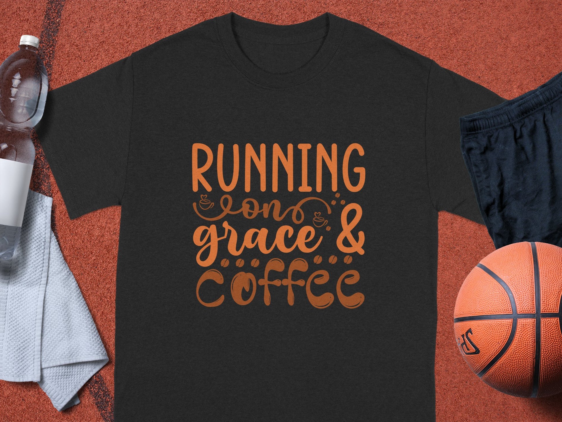 A Garment Graphics dark gray T-shirt with an orange Running on grace & coffee design sits on an orange surface surrounded by a basketball, black shorts, a white towel, and a half-filled water bottle—ideal for those who love Inspirational Sayings.