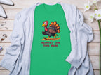 Garment Graphics offers a green T-shirt with a cartoon turkey and TURKEY ON THE RUN text, styled on wood beside a gray cardigan, jewelry, and white flowers. Ideal for those wanting funny graphic tees in family sizes.