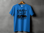 A blue Garment Graphics T-shirt is displayed on a wooden hanger against a gray wall. Featuring black text and graphics, it highlights Enjoy Classic Car with a vintage car image, celebrating the Vintage Ride Community since 1980 Aircooled. Ideal for car enthusiasts.
