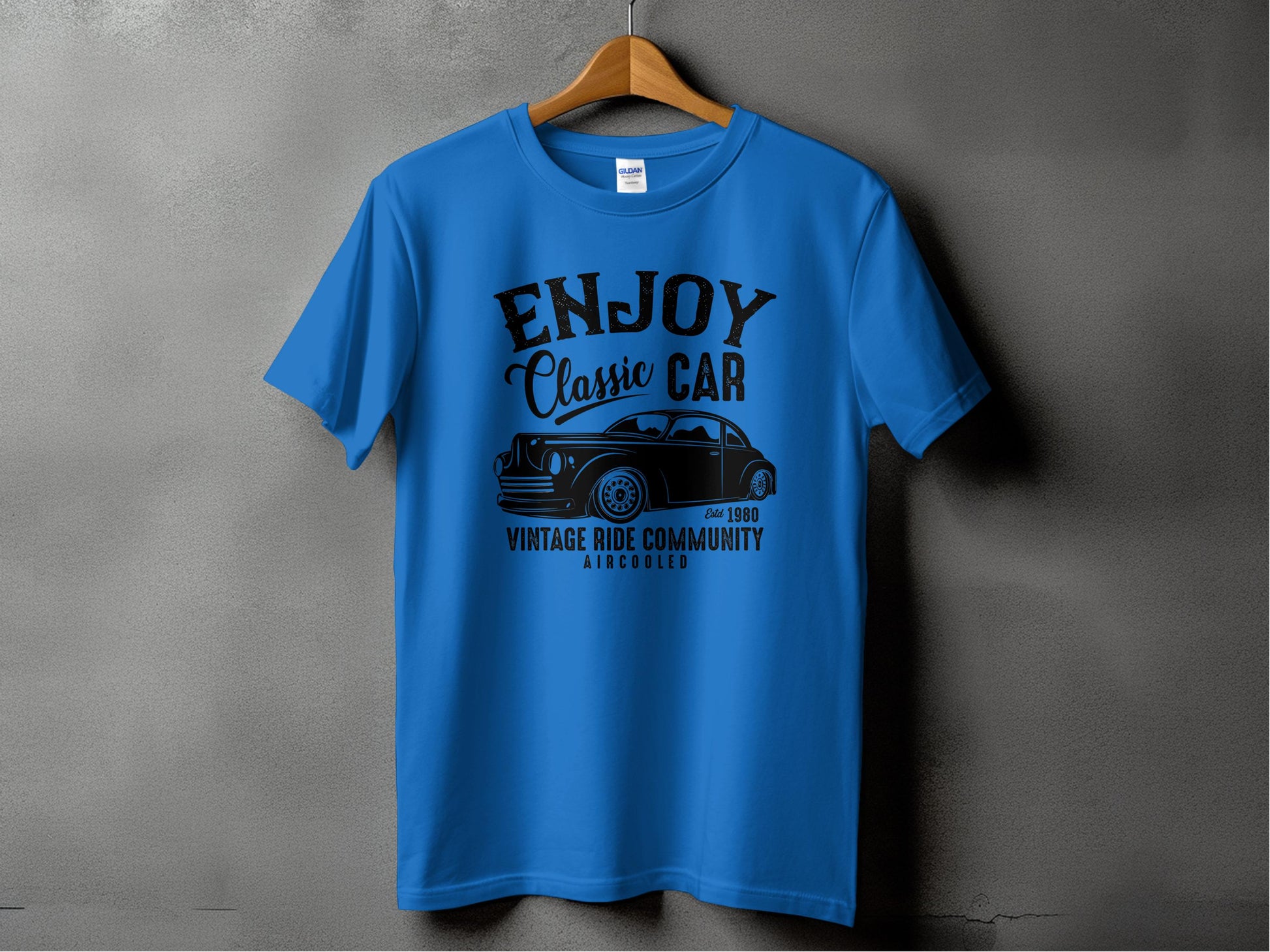 A blue Garment Graphics T-shirt is displayed on a wooden hanger against a gray wall. Featuring black text and graphics, it highlights Enjoy Classic Car with a vintage car image, celebrating the Vintage Ride Community since 1980 Aircooled. Ideal for car enthusiasts.