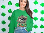 A smiling woman wears a Garment Graphics green graphic sweatshirt featuring an ornate eye design and the word GAZE. She stylishly pairs it with denim shorts, set against a backdrop of green clover-shaped decorations.