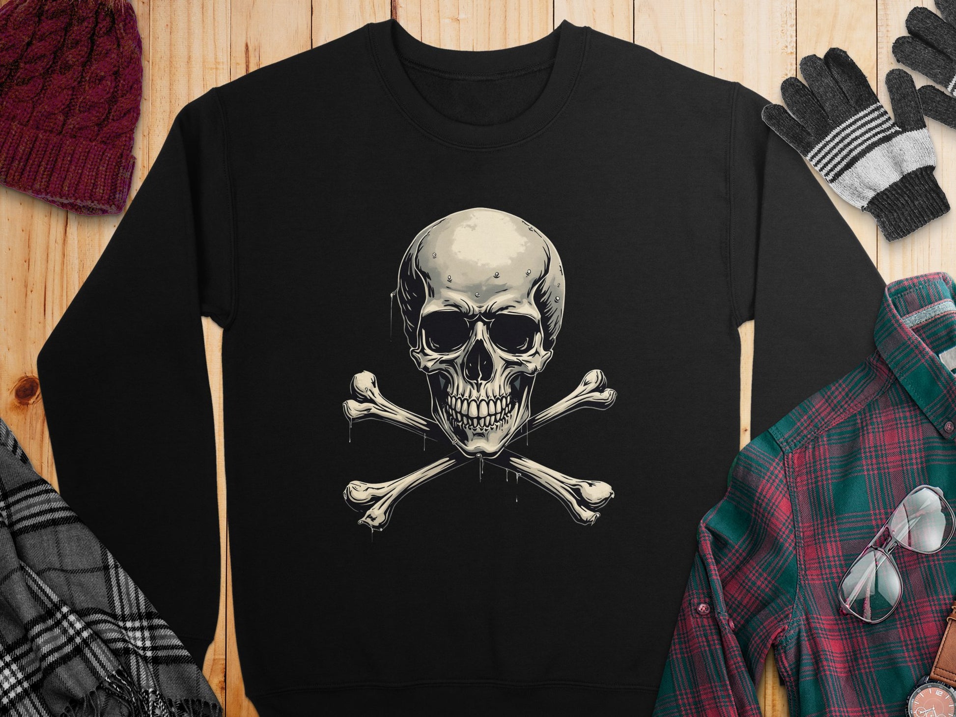 The Garment Graphics black sweatshirt, with an edgy skull and crossbones design, is displayed on wood. Its surrounded by a red knit hat, black and gray gloves, a plaid scarf, and a red and green plaid shirt—perfect pieces for its unisex appeal.