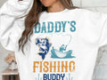 A person in a white shirt displaying Daddys Fishing Buddy by Garment Graphics stands with part of their face visible. The design is ideal for fishing lovers, featuring a fishing hook, fish, and someone casting from a boat.