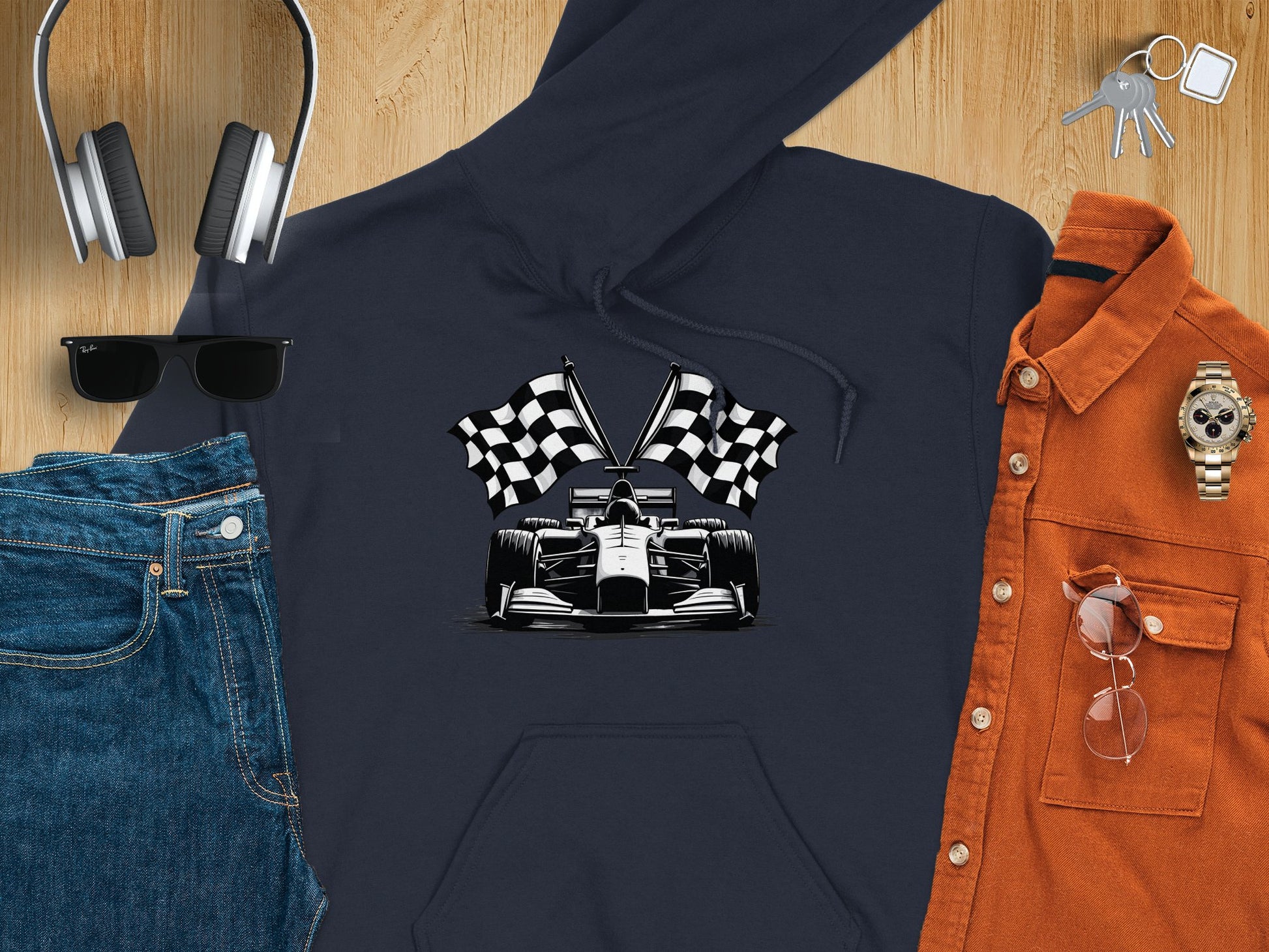 Garment Graphics flat lay showcases a trendy navy hoodie with checkered flag and race car print, paired with jeans, headphones, sunglasses, a brown button-up shirt, keys, a watch, and aviator sunglasses on wood—ideal for motorsport fans.