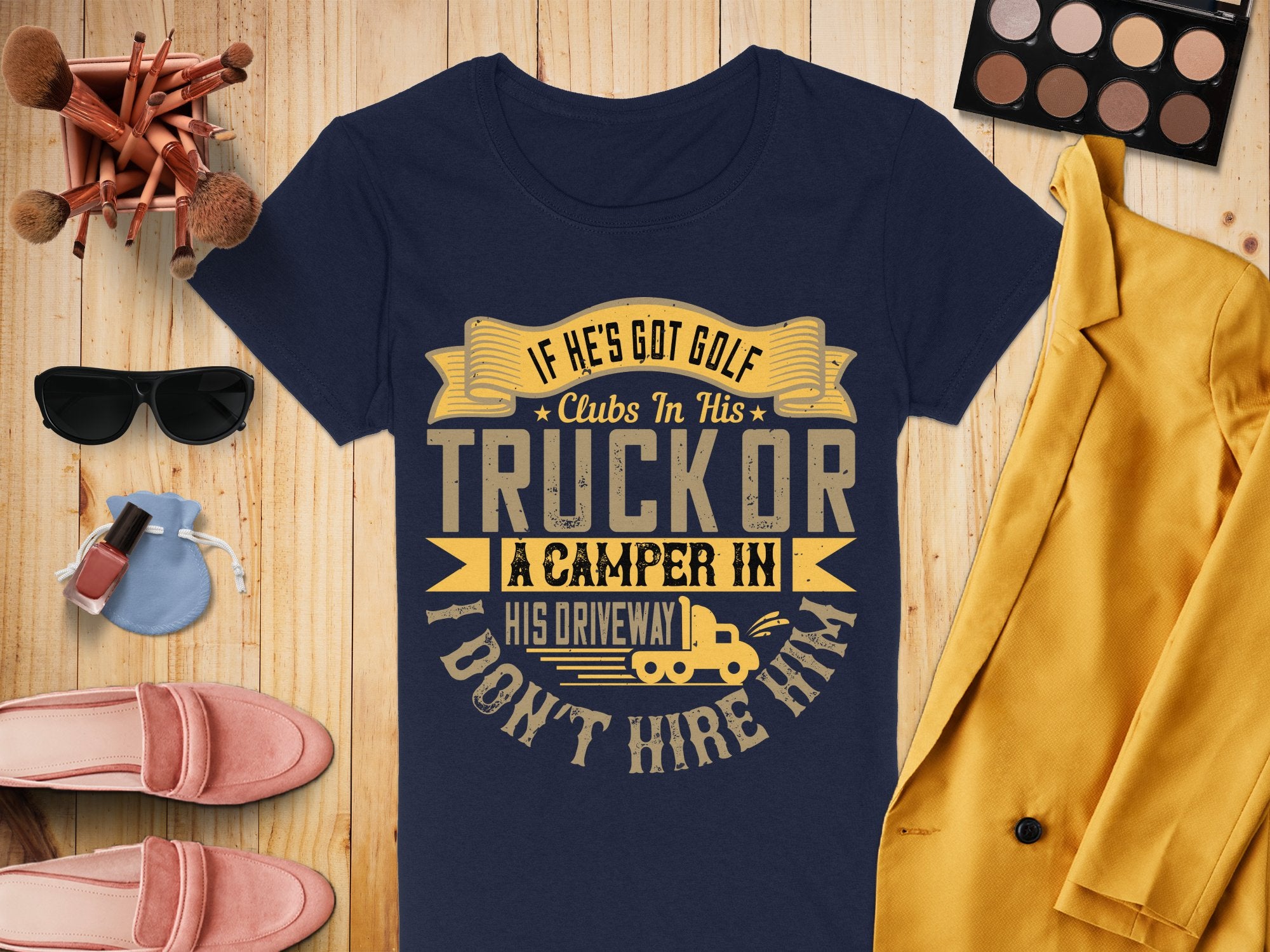 A flatlay features a quirky navy T-shirt from Garment Graphics with the funny golf quote, paired expertly with makeup brushes, an eyeshadow palette, a yellow coat, shoes, perfume, and a camera. Its available in womens sizes.