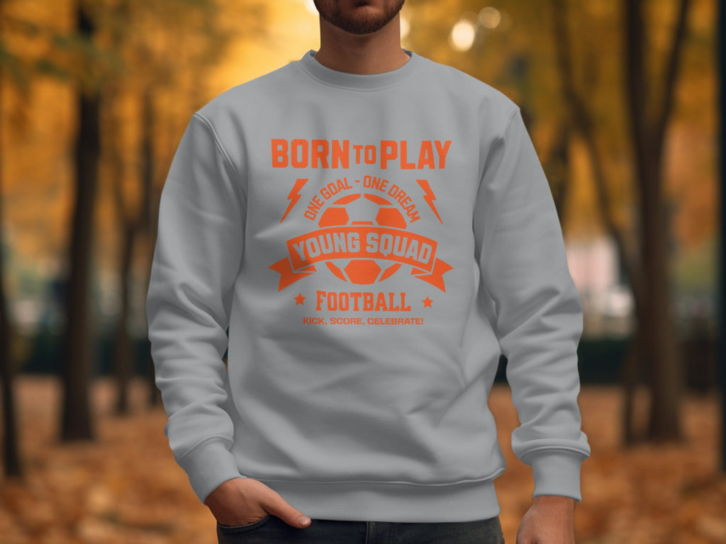 A person wears a Garment Graphics light gray football sweatshirt with Born to Play and Young Squad Football in orange text, perfect for young aspiring footballers. The autumn backdrop of trees and leaves suits the unisex sizing apparel perfectly.