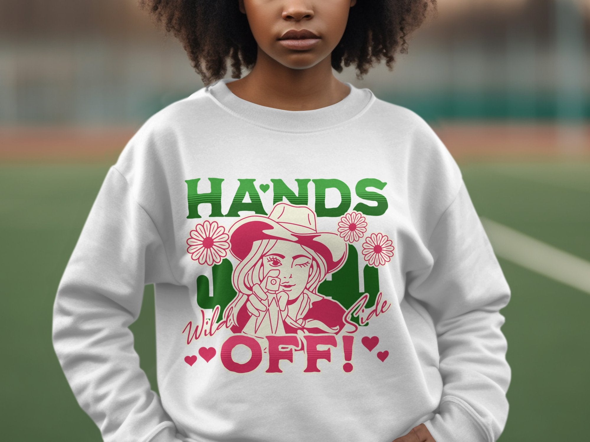 A person wears a white, loose-fit sweatshirt by Garment Graphics, featuring a colorful cowboy hat graphic adorned with flowers and HANDS OFF! Wild Side in green, pink, and red text, perfectly capturing the essence of their unique designs in an outdoor setting.
