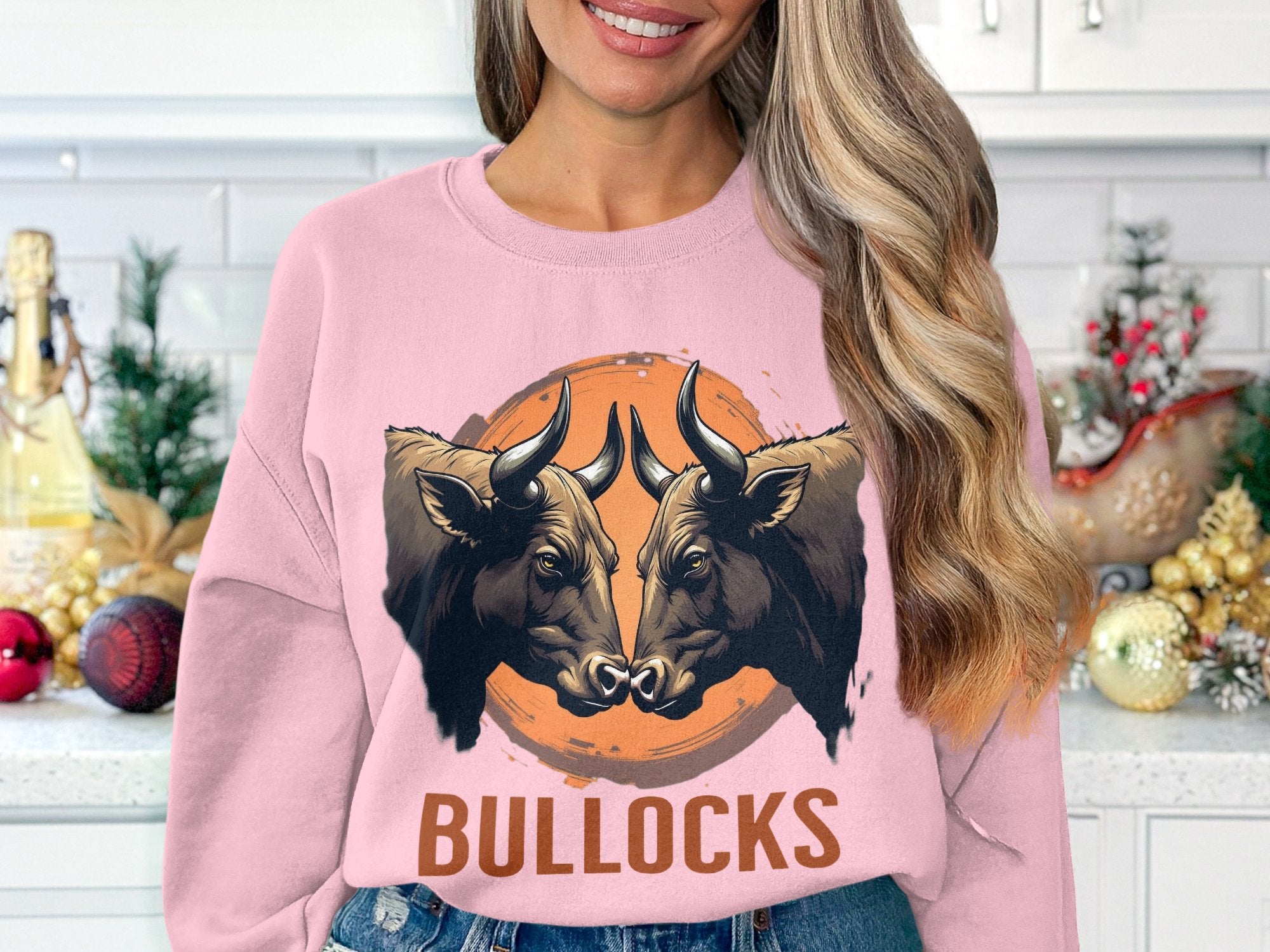 A person wears a loose fit pink sweatshirt by Garment Graphics, featuring a striking graphic of two bulls facing each other with BULLOCKS beneath. The kitchen in the background is festively decorated for the holidays.