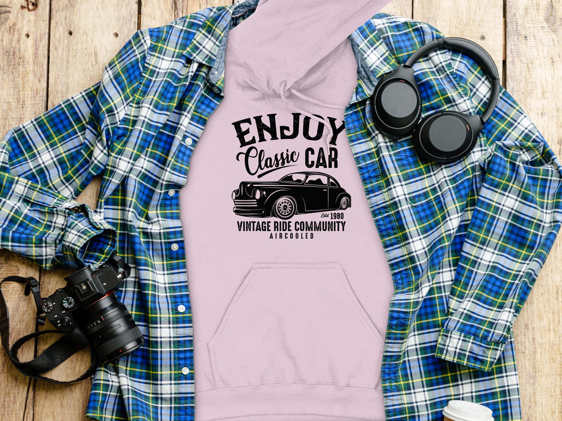 Flat lay of a lavender hoodie by Garment Graphics with a Classic Car graphic. Its paired with a blue plaid shirt, surrounded by black headphones, a camera, and a white cup with lid on a wooden surface.