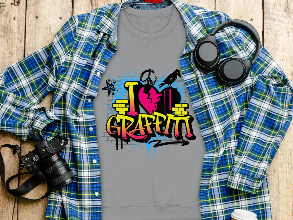 A gray Garment Graphics t-shirt featuring a lively I Love Graffiti design is layered under a blue plaid shirt. Nearby, black headphones, a strap-equipped camera, and a takeaway coffee cup on the wooden surface complete the casual street art vibe.