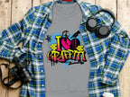 A gray Garment Graphics t-shirt featuring a lively I Love Graffiti design is layered under a blue plaid shirt. Nearby, black headphones, a strap-equipped camera, and a takeaway coffee cup on the wooden surface complete the casual street art vibe.