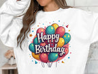 Someone is wearing a Garment Graphics white sweatshirt featuring colorful balloon designs and the text Happy Birthday. This unisex sweatshirt is perfect for celebrations, with a blurred white wall in the background.