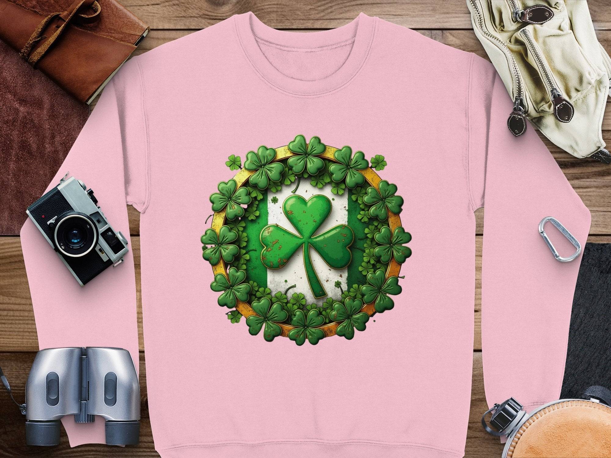 A pink sweatshirt from Garment Graphics rests on a wooden surface, featuring a lucky four-leaf clover encircled by smaller shamrocks. Surrounding this Irish-themed garment are accessories including a camera, binoculars, a leather pouch, and coiled rope.