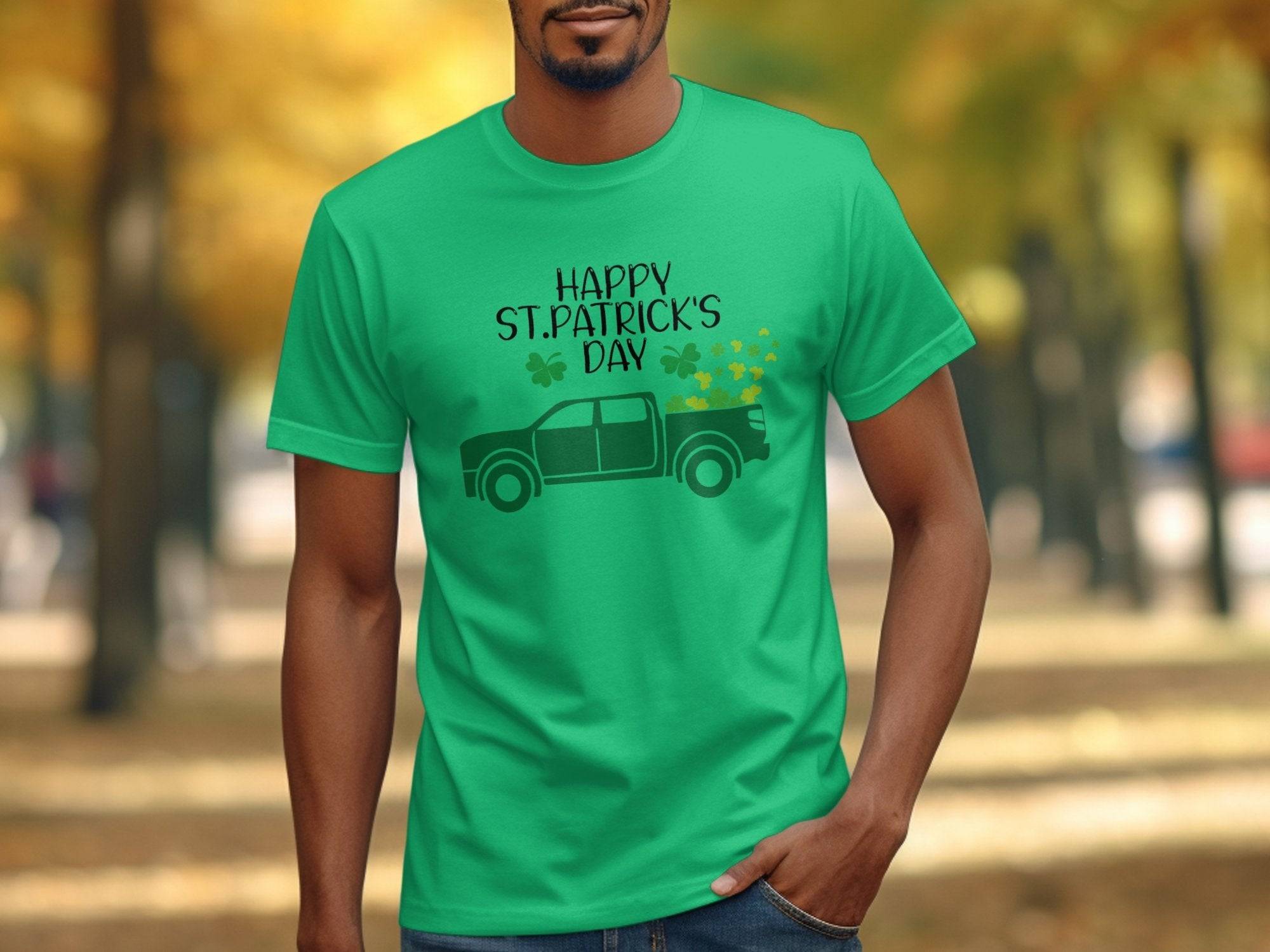Outside, a person wears a Garment Graphics green T-shirt with Happy St. Patricks Day, featuring shamrocks and a green pickup truck. Blurred trees with autumn leaves are in the background.