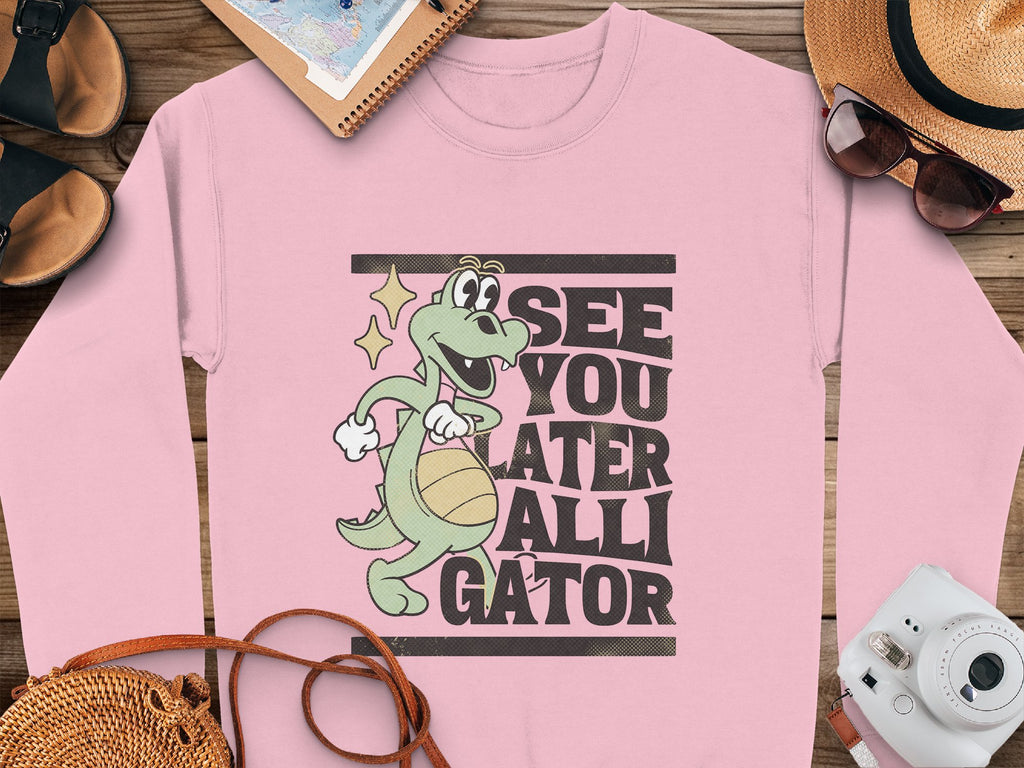 A pink Garment Graphics sweatshirt featuring a playful alligator with a basketball and the phrase See You Later Alligator rests on wood. Accessories like a straw hat, notebook, sunglasses, sandals, and a camera surround it.