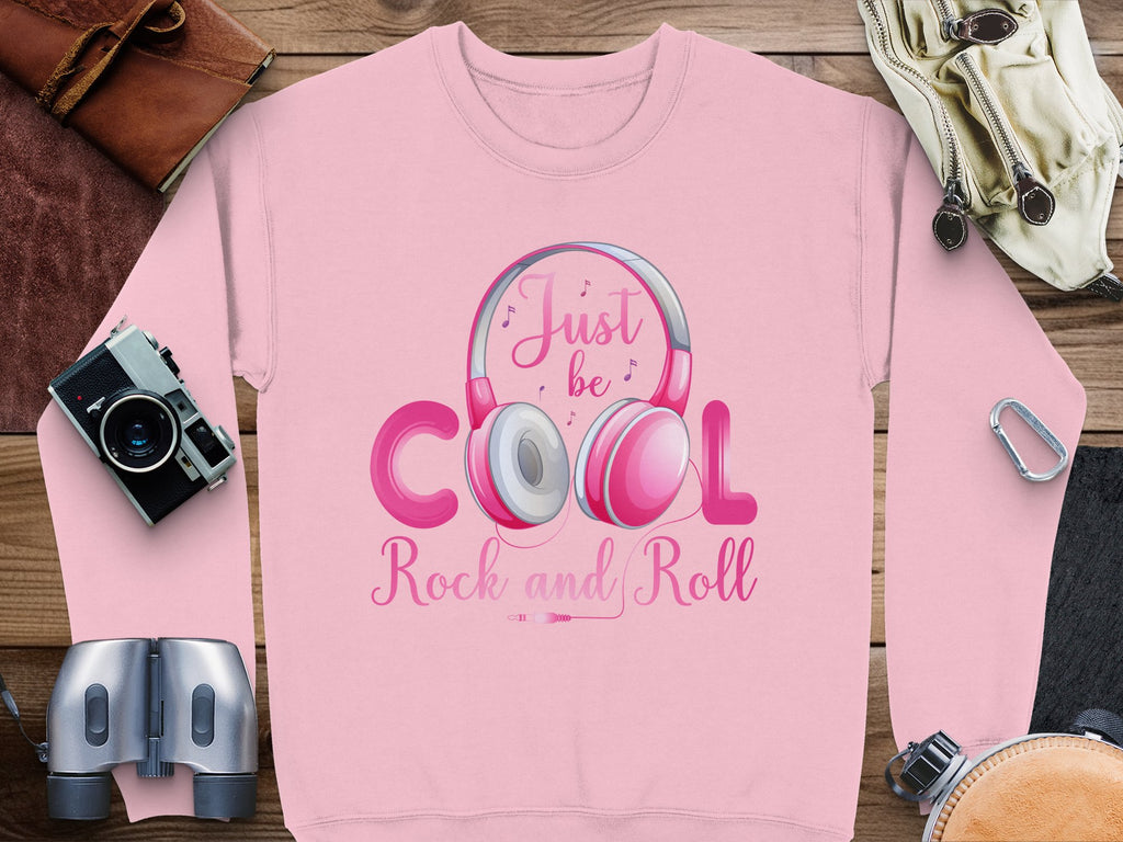 Garment Graphics pink sweatshirt showcases a Just be COOL Rock and Roll headphones graphic, ideal for music enthusiasts and fashion lovers. Its styled with a camera, notebook, binoculars, and leather accessories on a wooden surface.