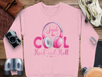 Garment Graphics pink sweatshirt showcases a Just be COOL Rock and Roll headphones graphic, ideal for music enthusiasts and fashion lovers. Its styled with a camera, notebook, binoculars, and leather accessories on a wooden surface.