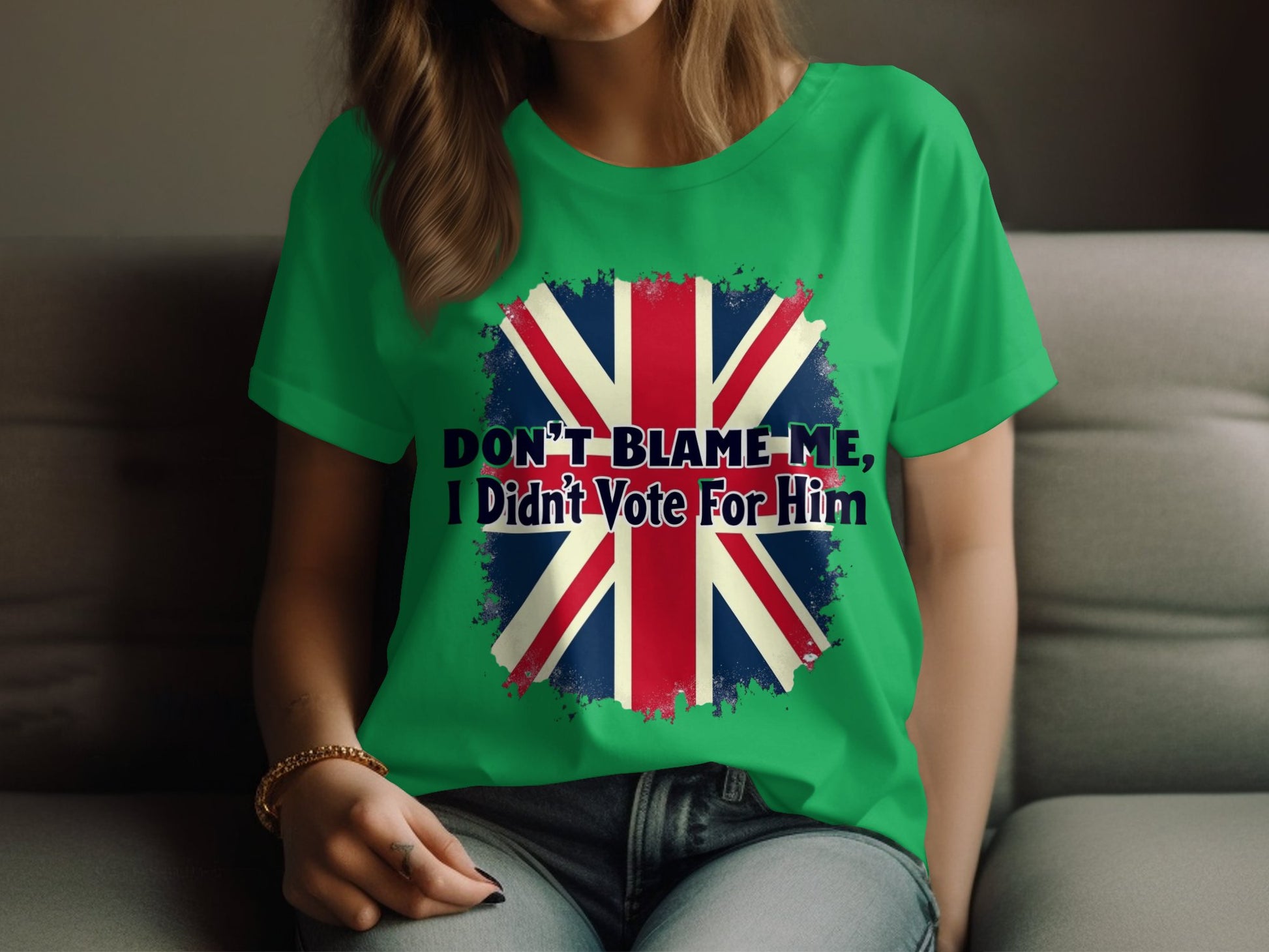 A person is lounging on a couch wearing a Garment Graphics green t-shirt made of 100% cotton. It features a striking distressed British flag and the bold text DONT BLAME ME, I Didnt Vote For Him.