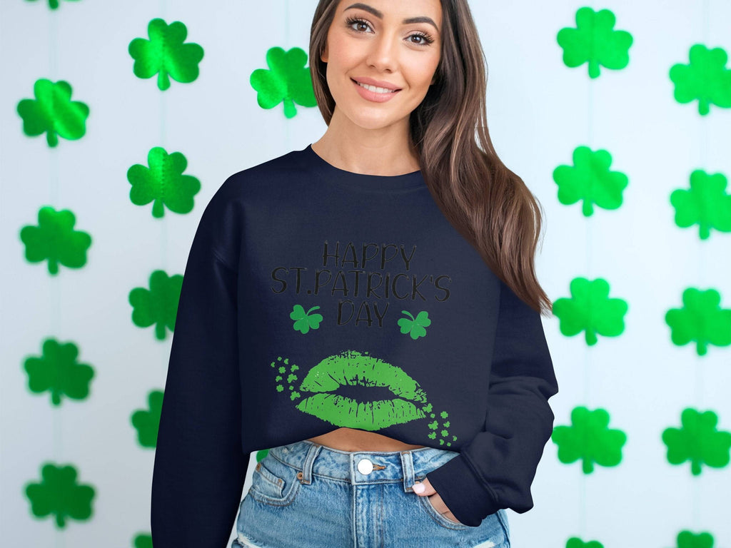 Someone stands before a white wall with green shamrocks, radiating festive spirit in a black Garment Graphics sweatshirt. The design features the phrase Happy St. Patricks Day with playful green lips, perfectly embodying a clover kiss theme.