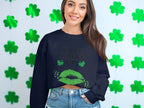 Someone stands before a white wall with green shamrocks, radiating festive spirit in a black Garment Graphics sweatshirt. The design features the phrase Happy St. Patricks Day with playful green lips, perfectly embodying a clover kiss theme.