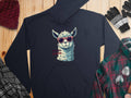 A Garment Graphics navy classic-fit hoodie with a llama in funny red sunglasses and No Prob-Llama text lies on wood, accompanied by a red beanie, striped gloves, plaid shirt, and black-and-white scarf.