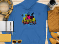 A blue Garment Graphics hoodie with an I Love Graffiti design is displayed on a wooden surface, ideal for casual wear and complements essentials like a notebook, pen, scarf, sunglasses, quilted purse, coffee cup, and mustard jacket.