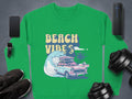 A Garment Graphics green sweatshirt with a Beach Vibes design, featuring a classic car, palm tree, and ocean wave. Its styled alongside a smartphone, smartwatch, black shoes, weights, and a portable speaker on a gray surface for that retro vibe.