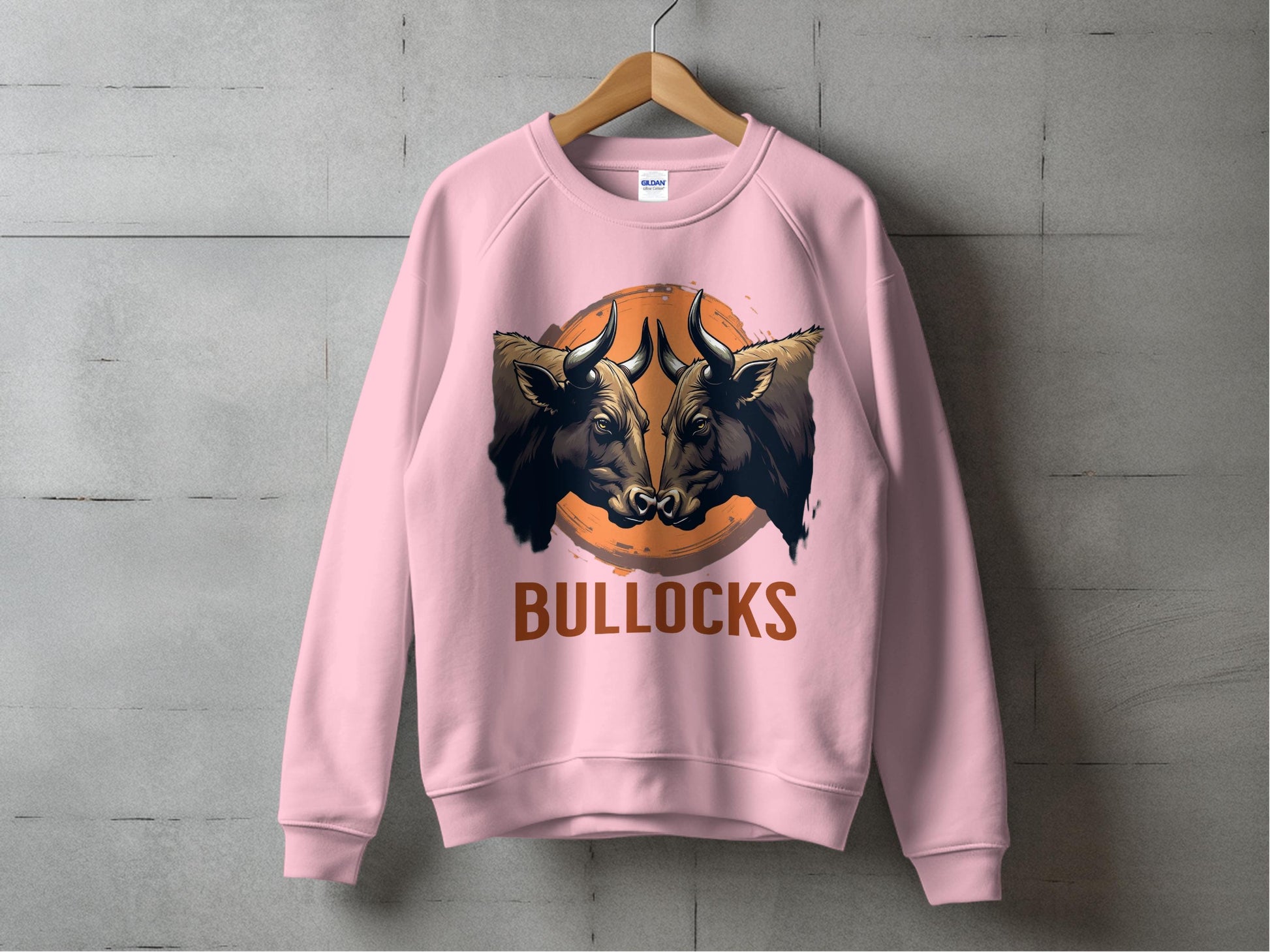 A pink unisex sweatshirt by Garment Graphics features two bold bulls facing forward above the stylized text BULLOCKS. Displayed on a wooden hanger against a concrete wall, this graphic sweatshirt suits everyone.