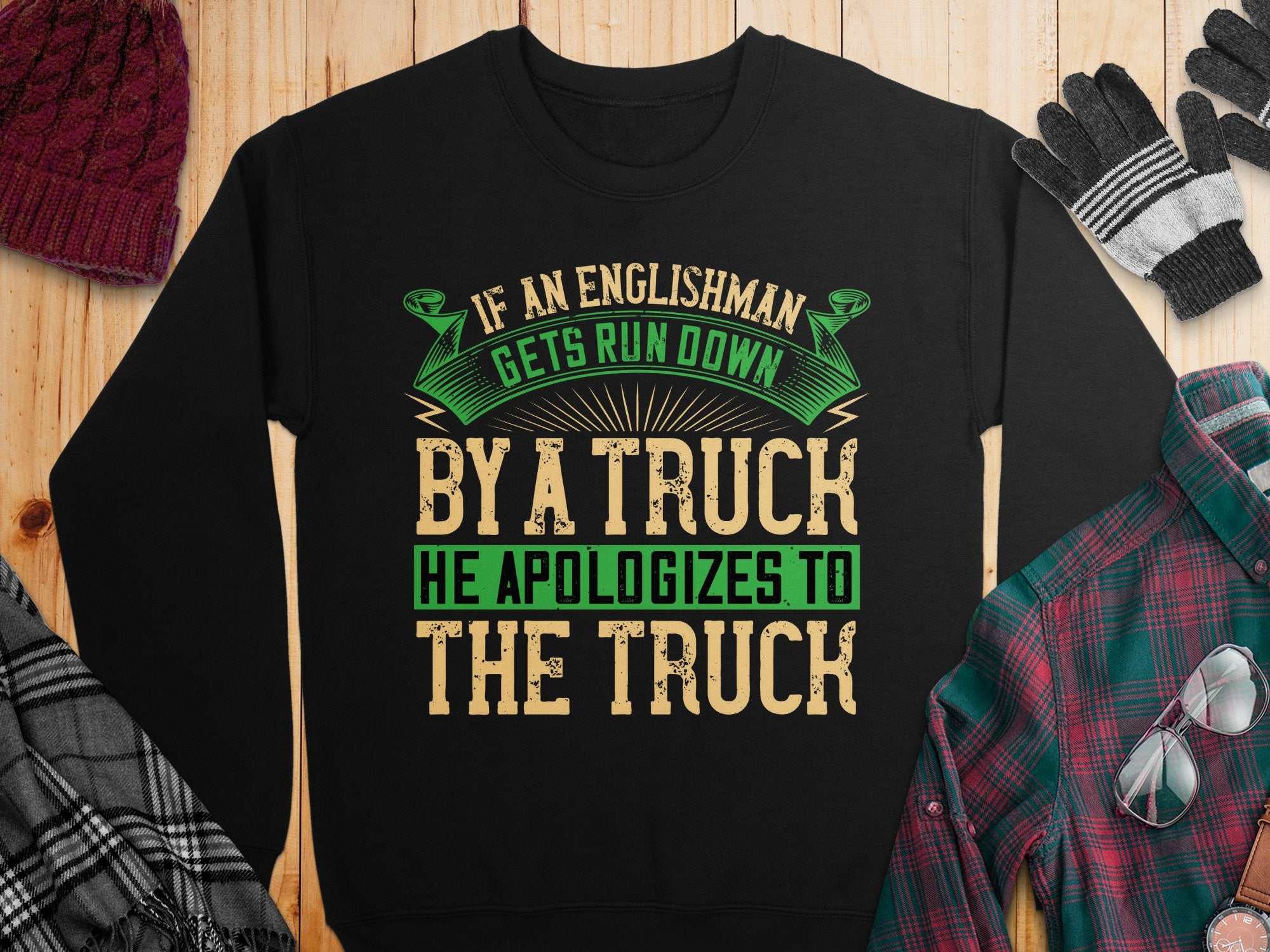 Garment Graphics black sweatshirt features a bold, humorous design with the phrase: If an Englishman gets run down by a truck he apologizes to the truck in green and beige letters. Its unisex-sized and styled on wood beside a red plaid shirt, gloves, and a red beanie.