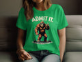 Indoors, someone wearing a Garment Graphics green T-shirt, adorned with Bigfoot making a peace sign and the quirky text ADMIT IT. Life would be boring without me, adds intrigue to any collection with this Admit It Bigfoot tee.