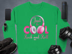 The Garment Graphics green sweatshirt with pink and white text, Just be Cool Rock and Roll, is ideal for music lovers. It showcases a headphone graphic surrounded by a smartphone, smartwatch, portable speaker, black sneakers, and adjustable dumbbells on a gray background.