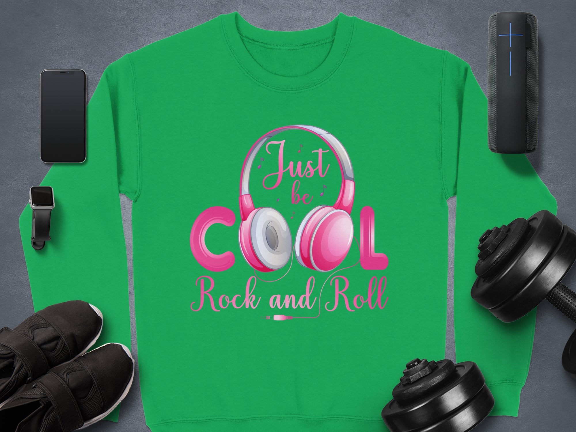The Garment Graphics green sweatshirt with pink and white text, Just be Cool Rock and Roll, is ideal for music lovers. It showcases a headphone graphic surrounded by a smartphone, smartwatch, portable speaker, black sneakers, and adjustable dumbbells on a gray background.