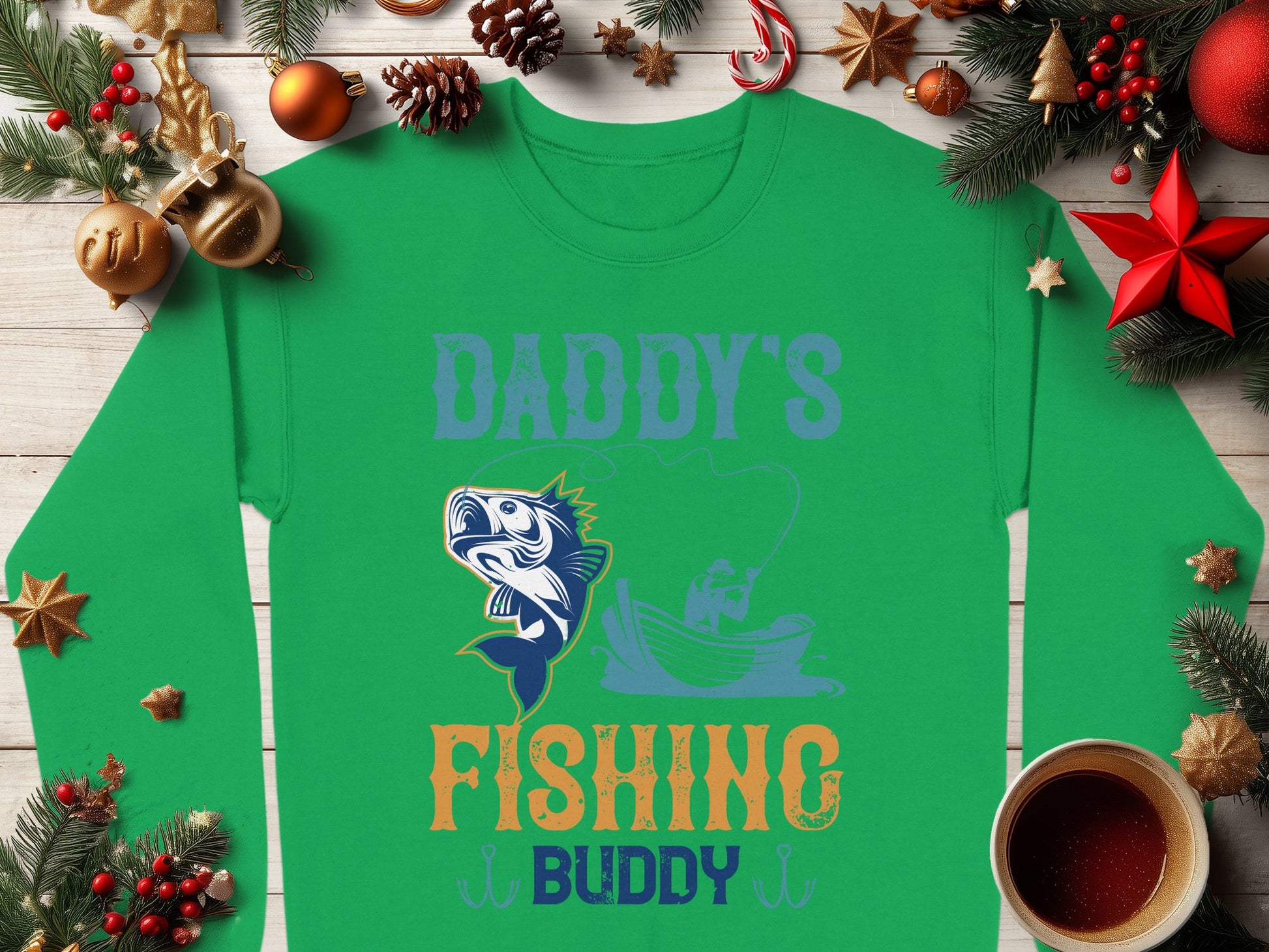 A green Garment Graphics shirt with a playful Daddys Fishing Buddy design sits on a wooden table decorated with pine branches, ornaments, and a steaming cup of coffee—ideal for outdoor enthusiasts who love graphic sweatshirts.