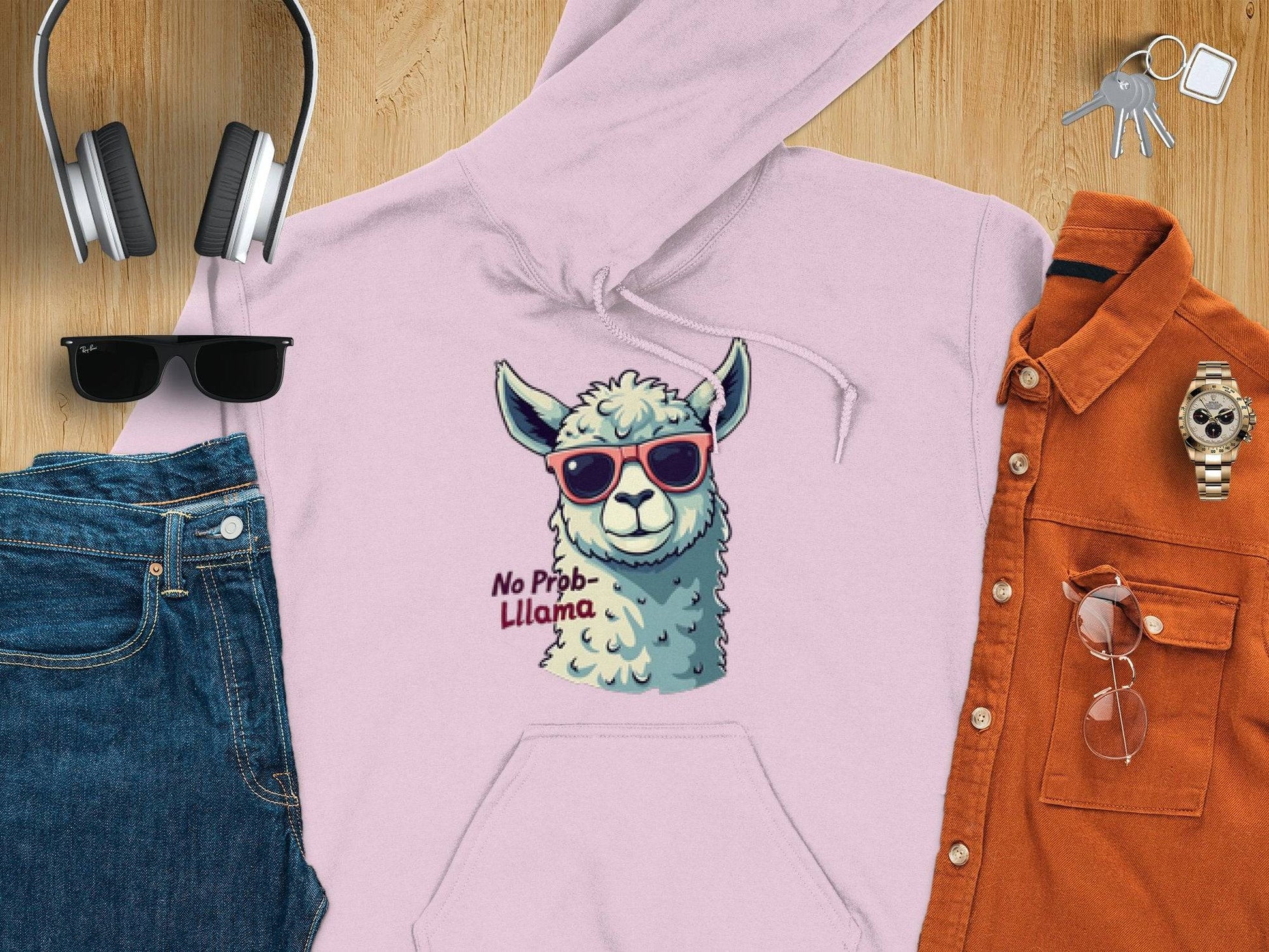 A flat lay image from Garment Graphics features a classic fit pink hoodie with a llama in funny sunglasses and the text No Prob-Llama, paired with blue jeans, black sunglasses, headphones, keys, a wristwatch, and an orange shirt with glasses.