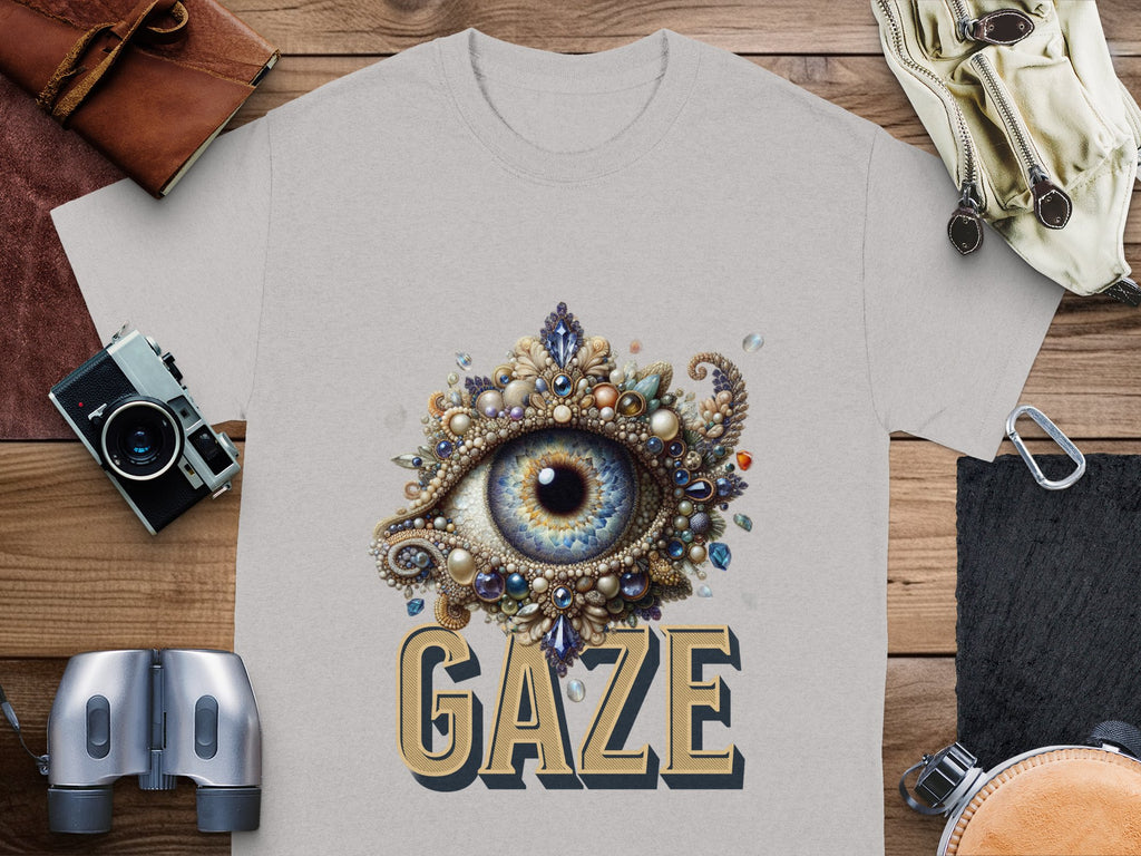 A gray graphic T-shirt by Garment Graphics features an artistic eye gaze design with ornate patterns and gemstones. GAZE is boldly inscribed beneath. This classic fit shirt is showcased on a wooden surface with various accessories around it.