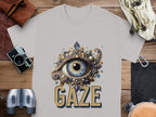 A gray graphic T-shirt by Garment Graphics features an artistic eye gaze design with ornate patterns and gemstones. GAZE is boldly inscribed beneath. This classic fit shirt is showcased on a wooden surface with various accessories around it.