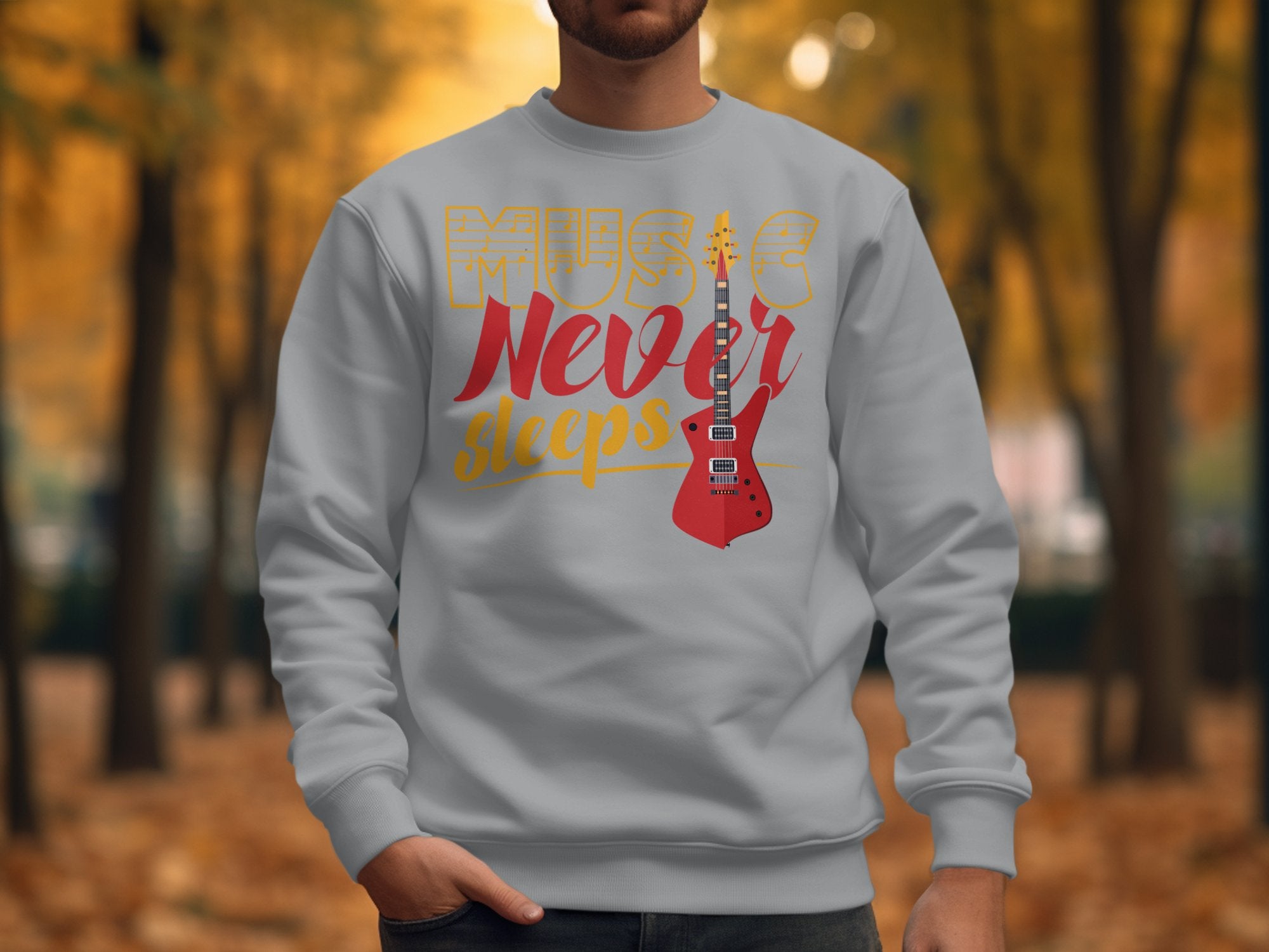 A person wearing a Garment Graphics light gray sweatshirt featuring a bold Music Never Sleeps print and a red electric guitar graphic, perfect for music lovers. The background shows an artistically blurred autumn scene with orange and yellow leaves.