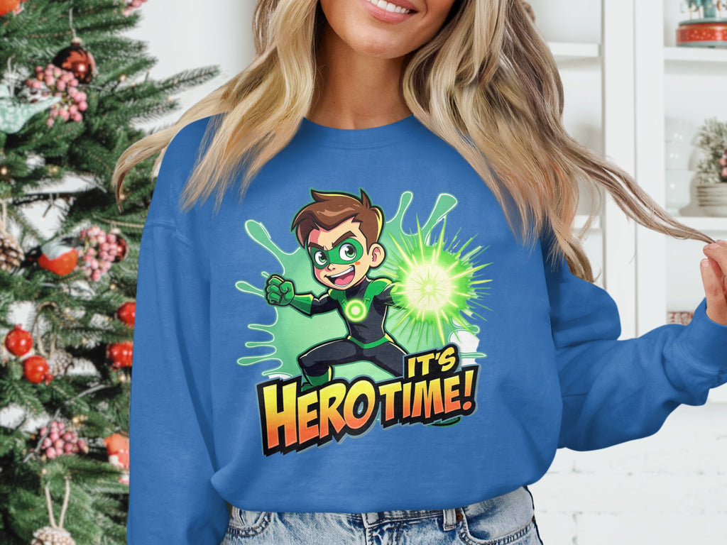 A person wears a blue graphic print sweatshirt by Garment Graphics featuring a superhero with brown hair in a green suit, the words Its Hero Time! and is surrounded by a glowing burst. A softly twinkling Christmas tree in the background adds festive charm.