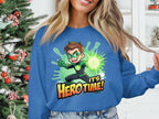 A person wears a blue graphic print sweatshirt by Garment Graphics featuring a superhero with brown hair in a green suit, the words Its Hero Time! and is surrounded by a glowing burst. A softly twinkling Christmas tree in the background adds festive charm.
