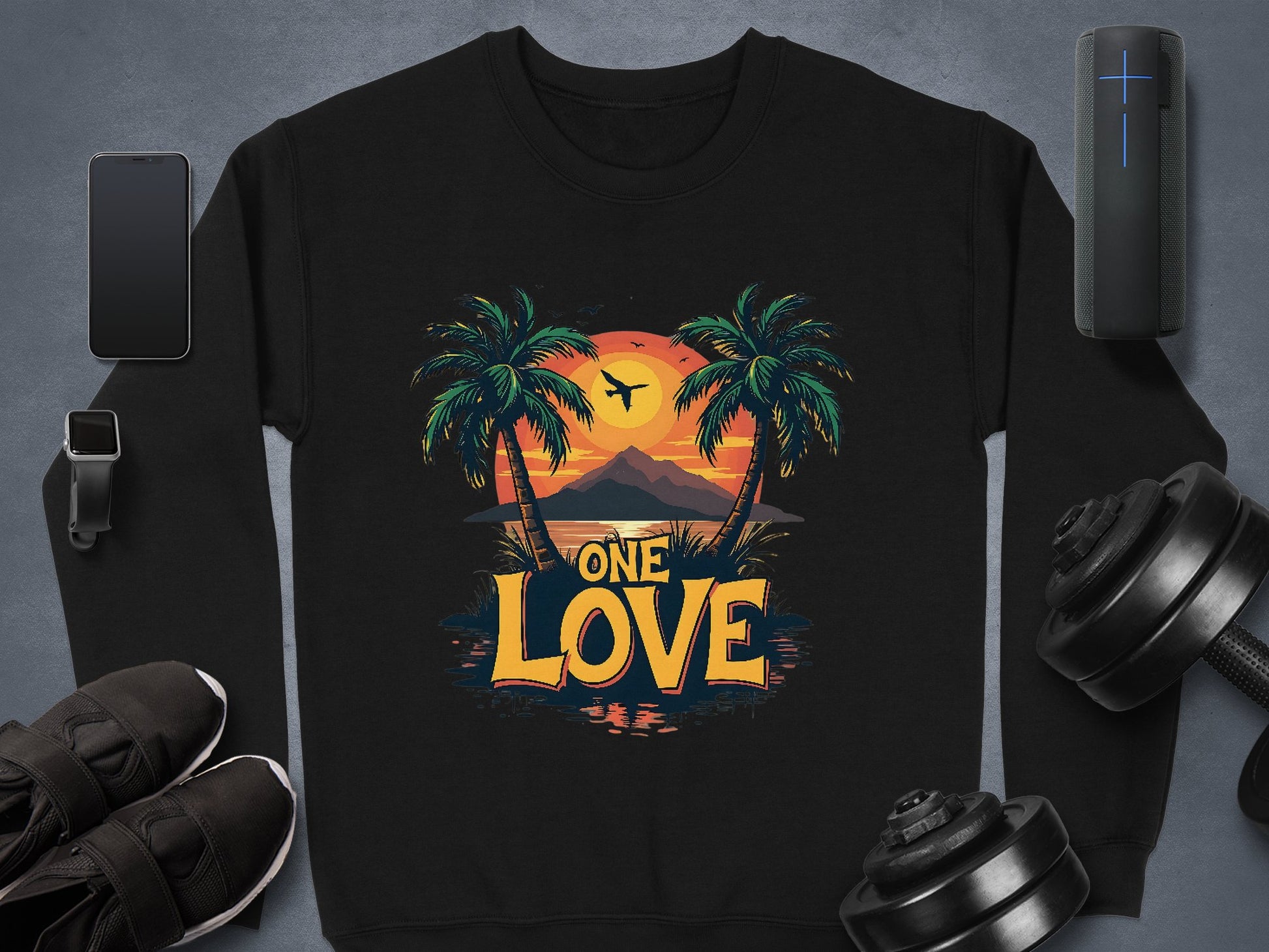 Garment Graphics black sweatshirt from the One Love Sweatshirts collection features a tropical sunset with palm trees, an airplane, and mountains. Unisex sizing ensures a perfect fit for all. Displayed with smartphone, smartwatch, earbuds, athletic shoes, and dumbbells on a gray surface.