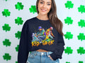 A woman beams in her Garment Graphics black sweatshirt, featuring a vibrant graphic of two people with Risk Taker Street Dance. She confidently poses before a wall of green shamrocks, embodying street dance culture.