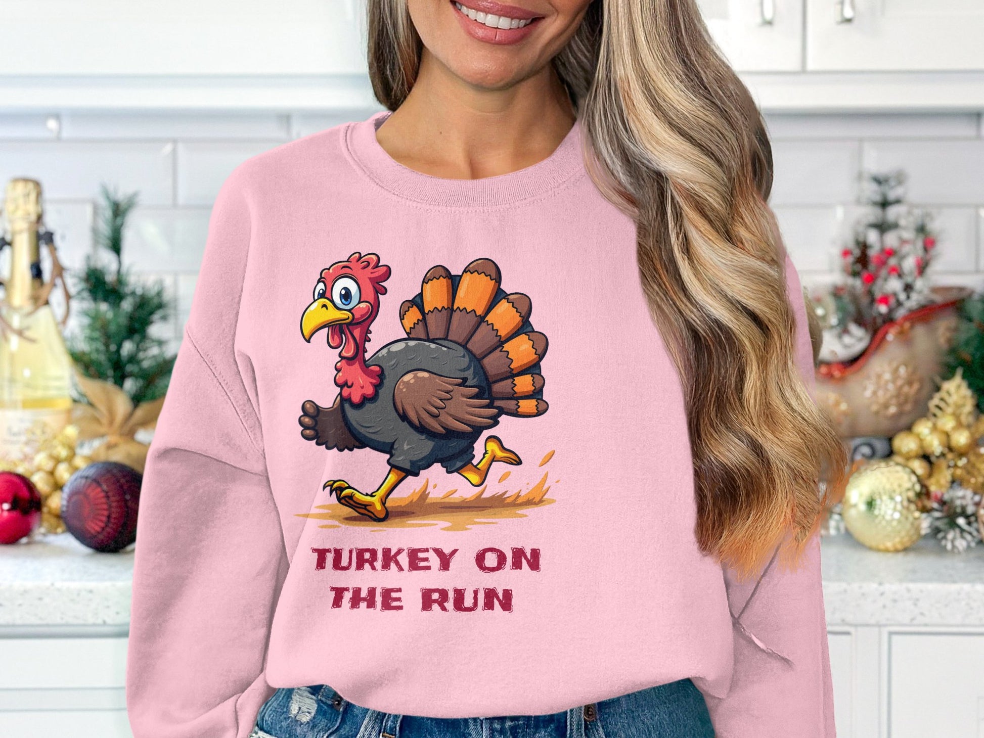 A woman in a pink Garment Graphics unisex sweatshirt with a cartoon turkey and Turkey on the Run stands in a festively decorated kitchen with small Christmas trees and ornaments.