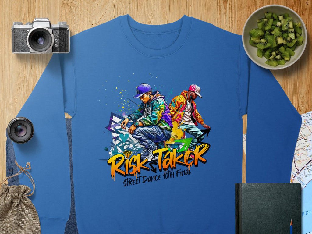 Garment Graphics blue sweatshirt showcases two street dancers in vibrant outfits with graffiti accents and Risk Taker Street Dance Finals text. Nearby are a camera, cucumber bowl, lenses, map, bag, and book. Ideal for anyone embracing street dances bold energy.
