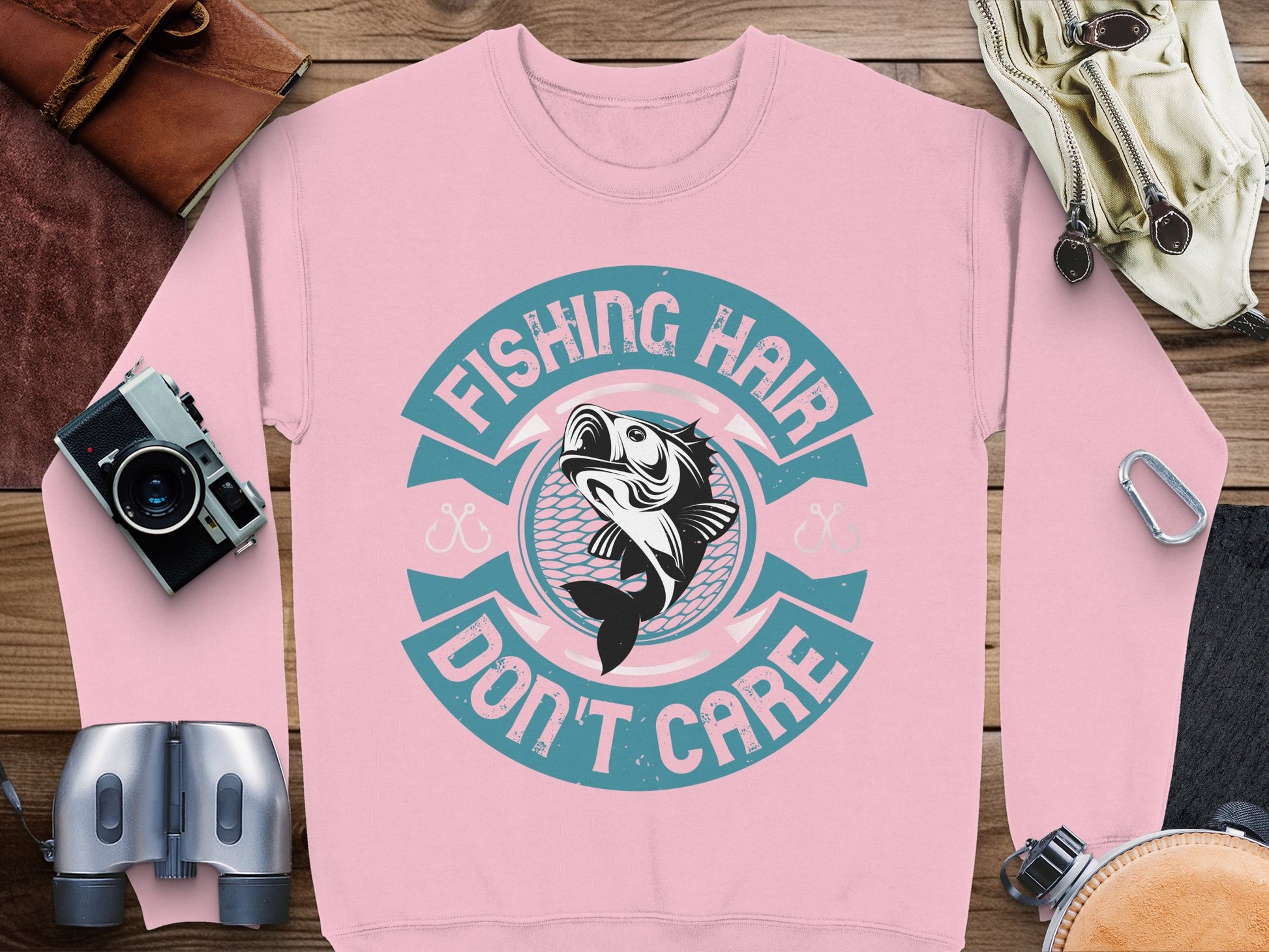 The pink fishing sweatshirt from Garment Graphics features the slogan Fishing Hair Dont Care and a playful fish graphic. Its perfect for any fishing enthusiast and is showcased with a camera, binoculars, a belt, a bag, and folded leather on a wooden surface.