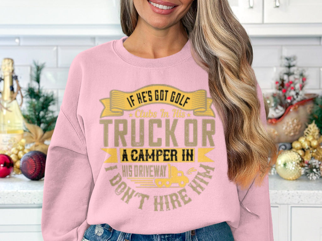 In a kitchen filled with festive decor, a woman wears Garment Graphics pink sweatshirt featuring the humorous slogan, If hes got golf clubs in his truck or a camper in his driveway I dont hire him.