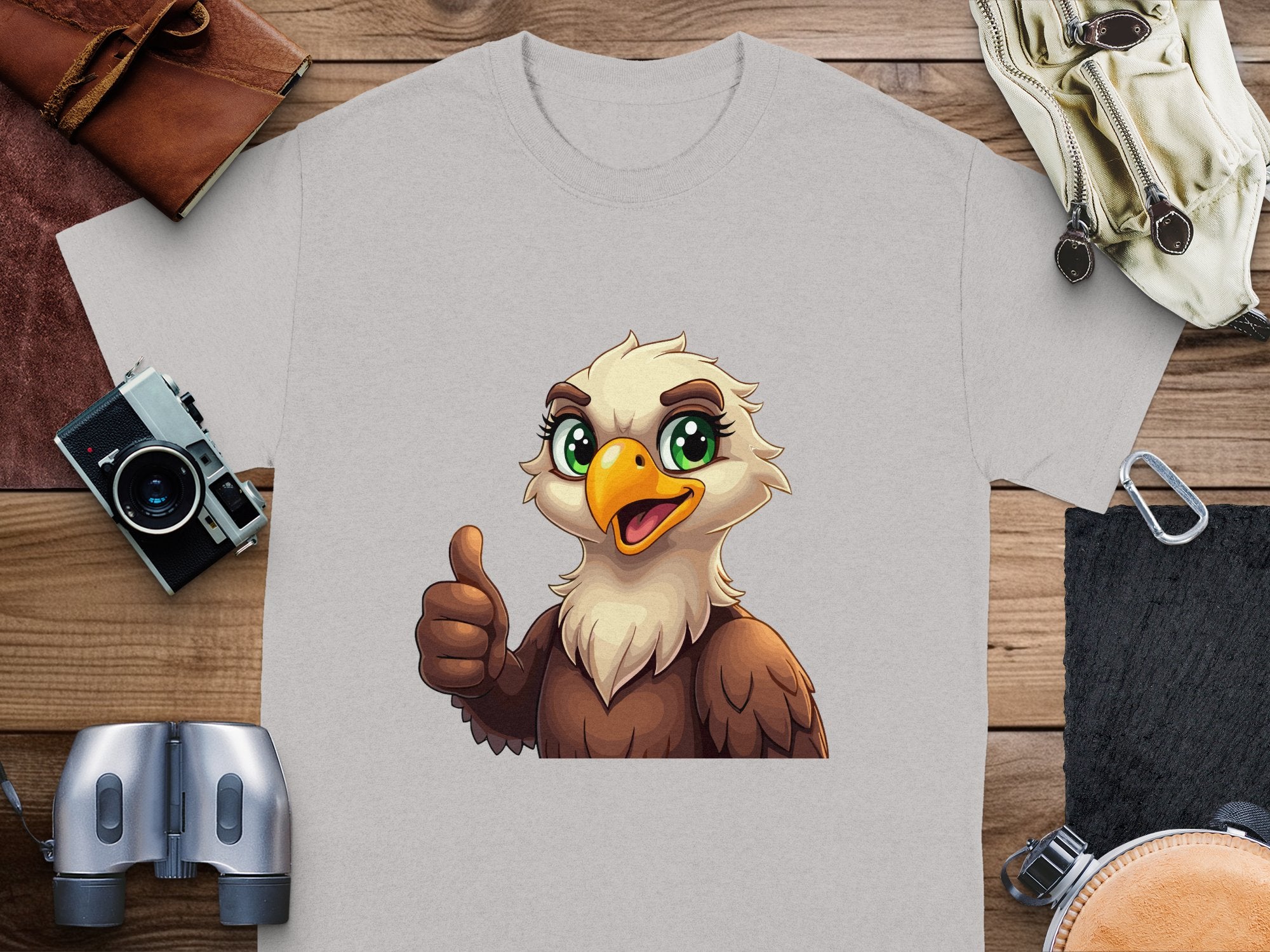 A light gray Garment Graphics T-shirt with a cartoon eagle giving a thumbs-up sits on a wooden surface. Ideal for a fun casual wardrobe, it pairs well with the leather wallet, vintage camera, binoculars, and gloves around it.